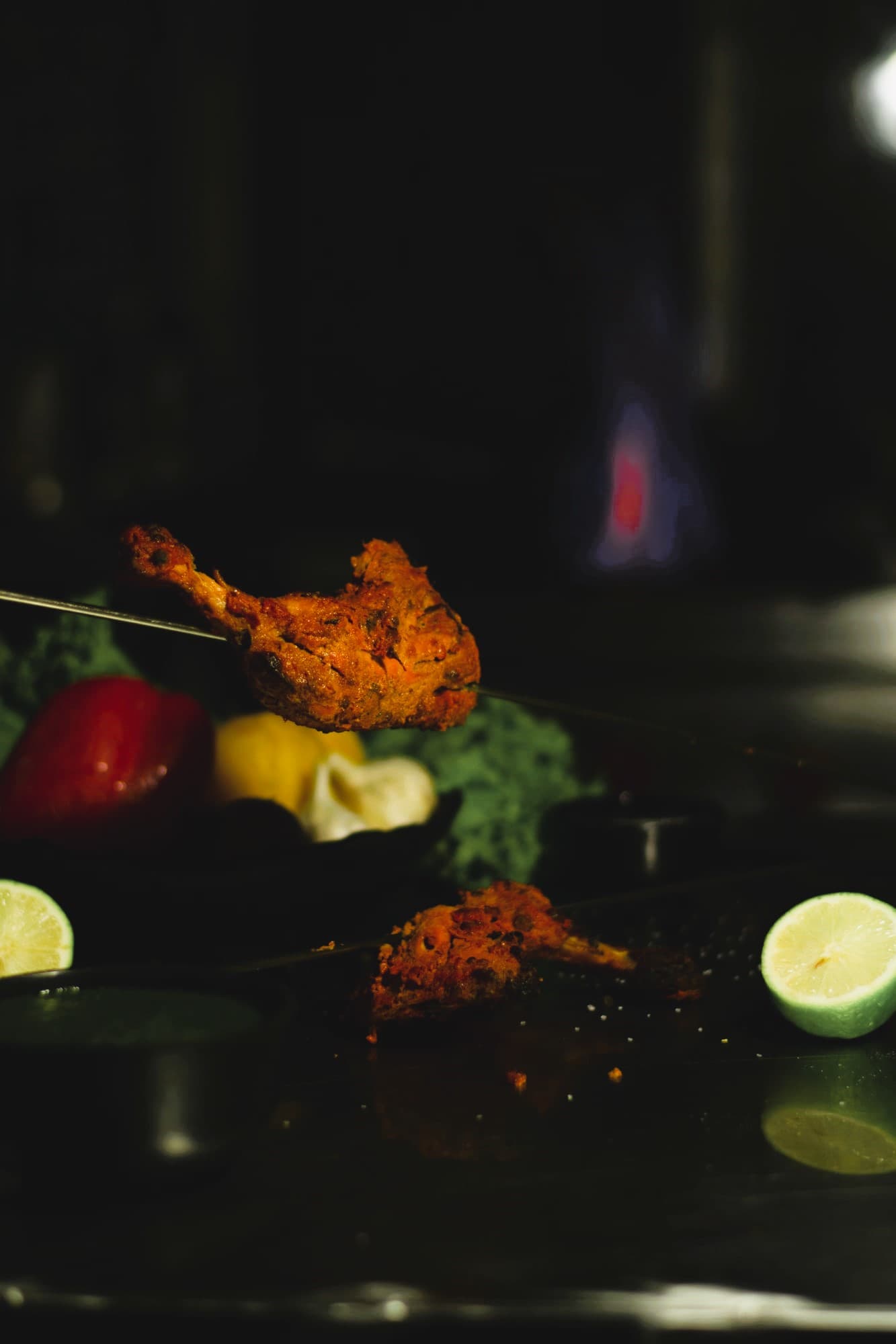 A single spiced drumstick on a steel skewer held over a dark grill, a blue gas flame burning behind it, lime halves and a cup of green sauce below.