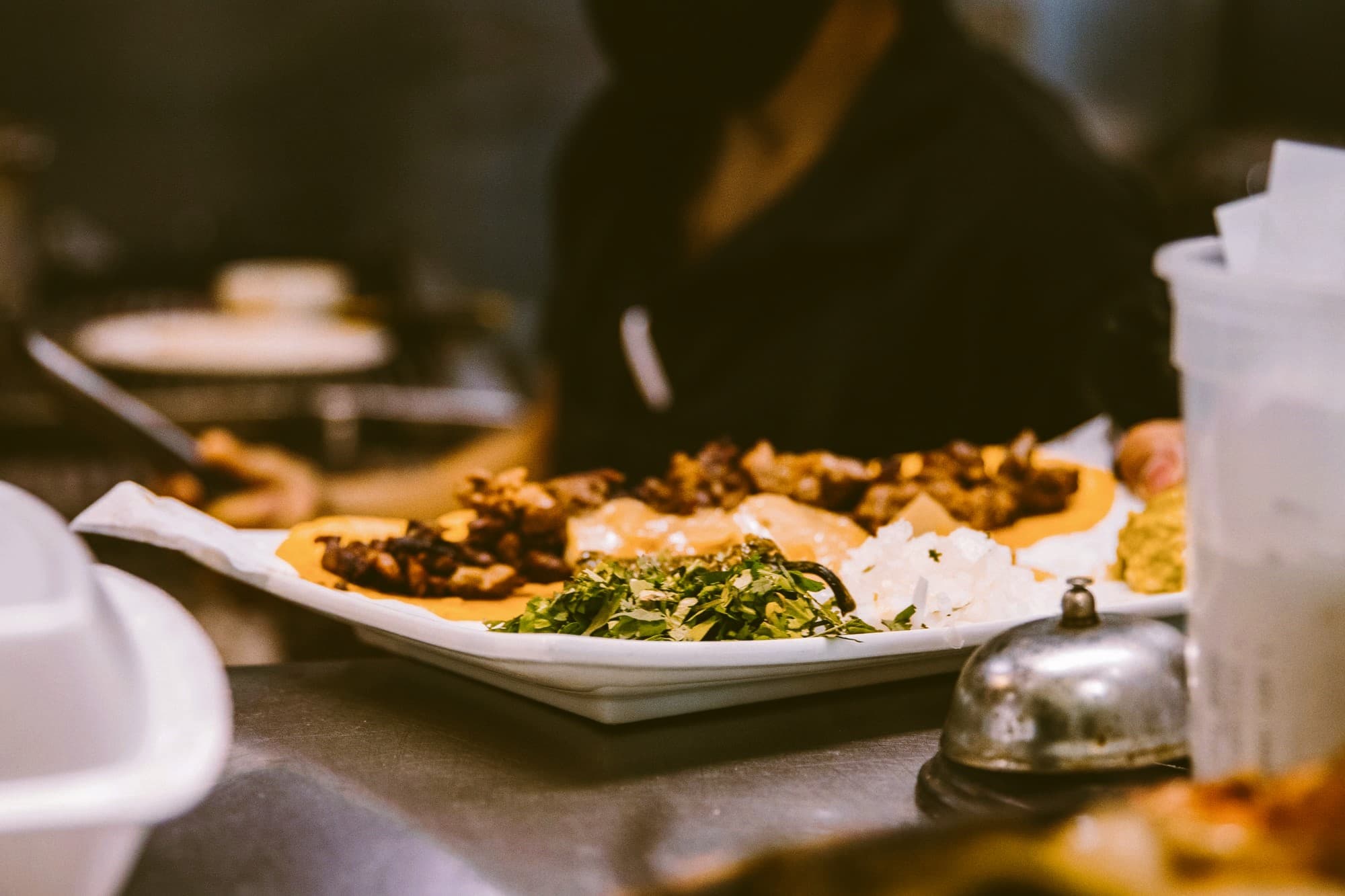 A long white plate on the kitchen pass holding chopped suya, boiled yam in sauce, white rice and a mound of chopped herbs, a cook working behind it.